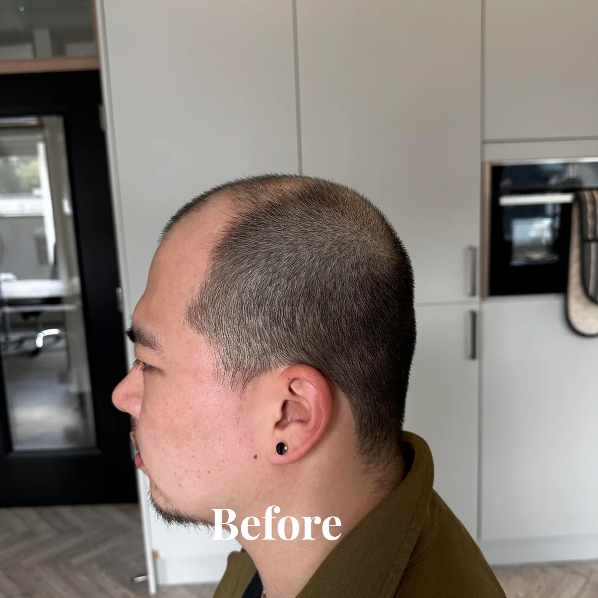 Side profile of a man with short buzz-cut hair and a trimmed mustache and goatee, wearing a black stud earring and an olive green jacket, with kitchen cabinets in the background and the word 'Before' overlayed.