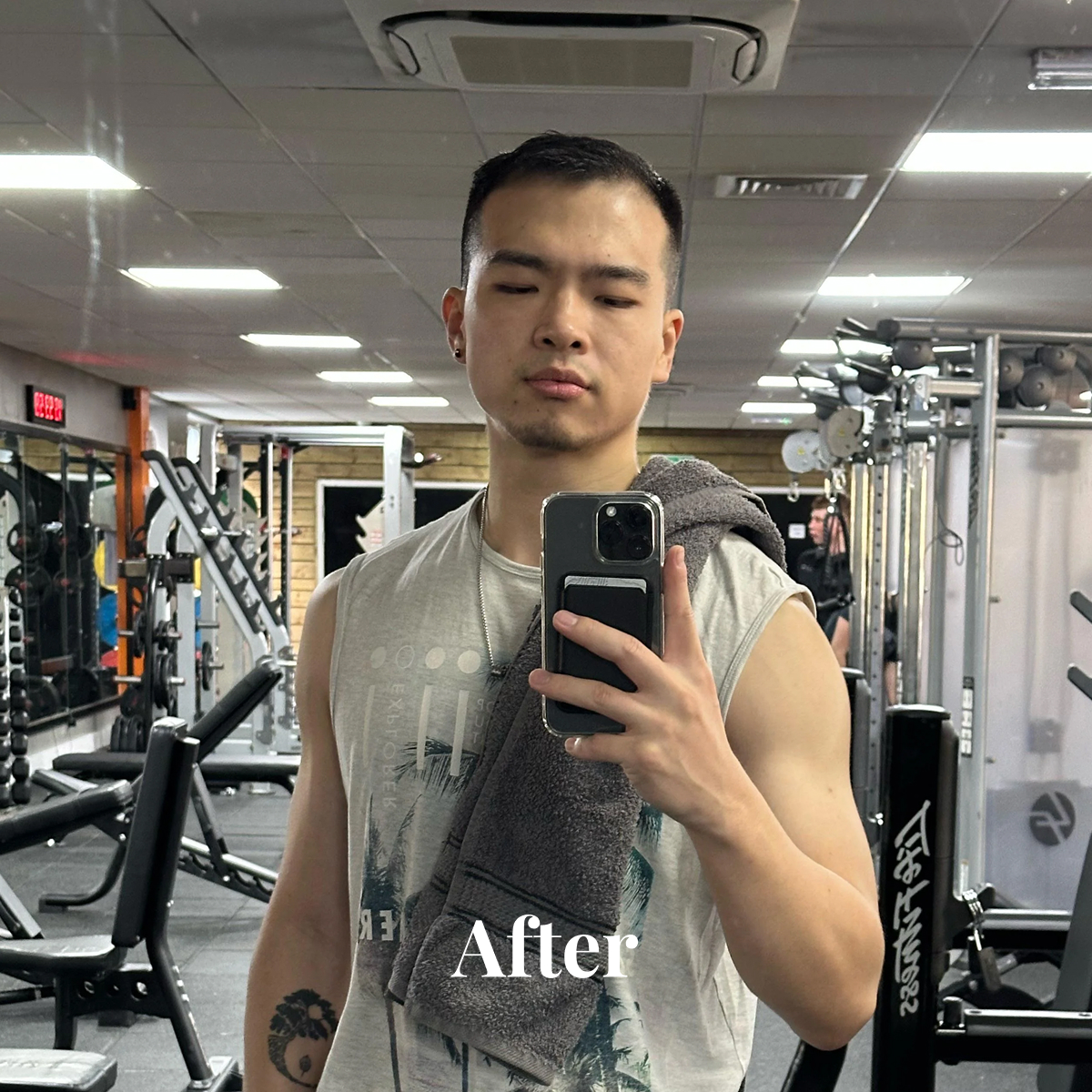 Young man taking a gym mirror selfie with a towel over his shoulder, wearing a sleeveless shirt.