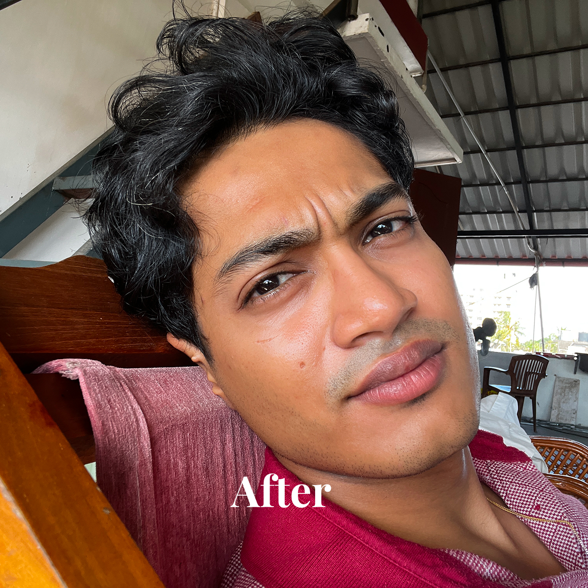 Close-up of a young man with short curly dark hair leaning against a wooden chair with a pink cloth, wearing a red checkered shirt.