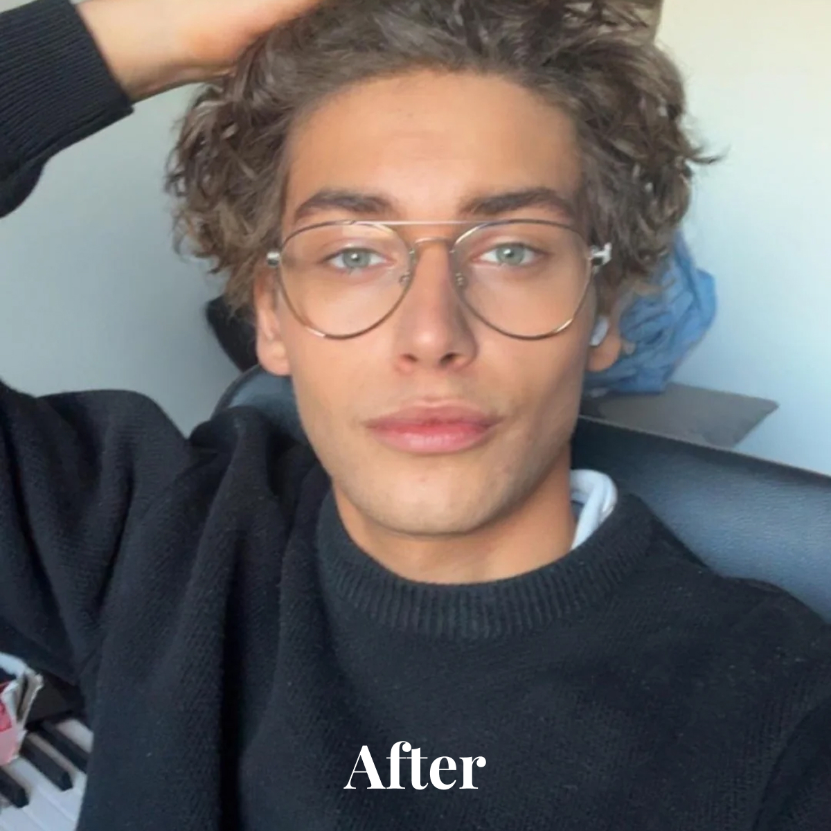 Young person with curly hair wearing large glasses and a black sweater posing indoors with one arm raised.