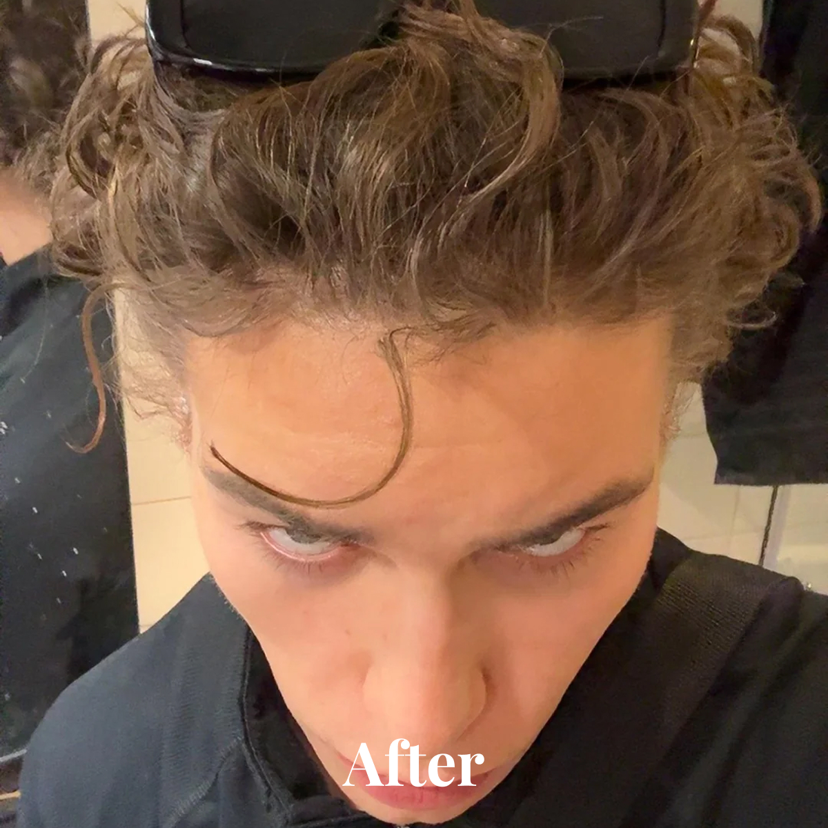 Top-down view of a person with curly brown hair and a prominent curl falling on their forehead, wearing a black garment.