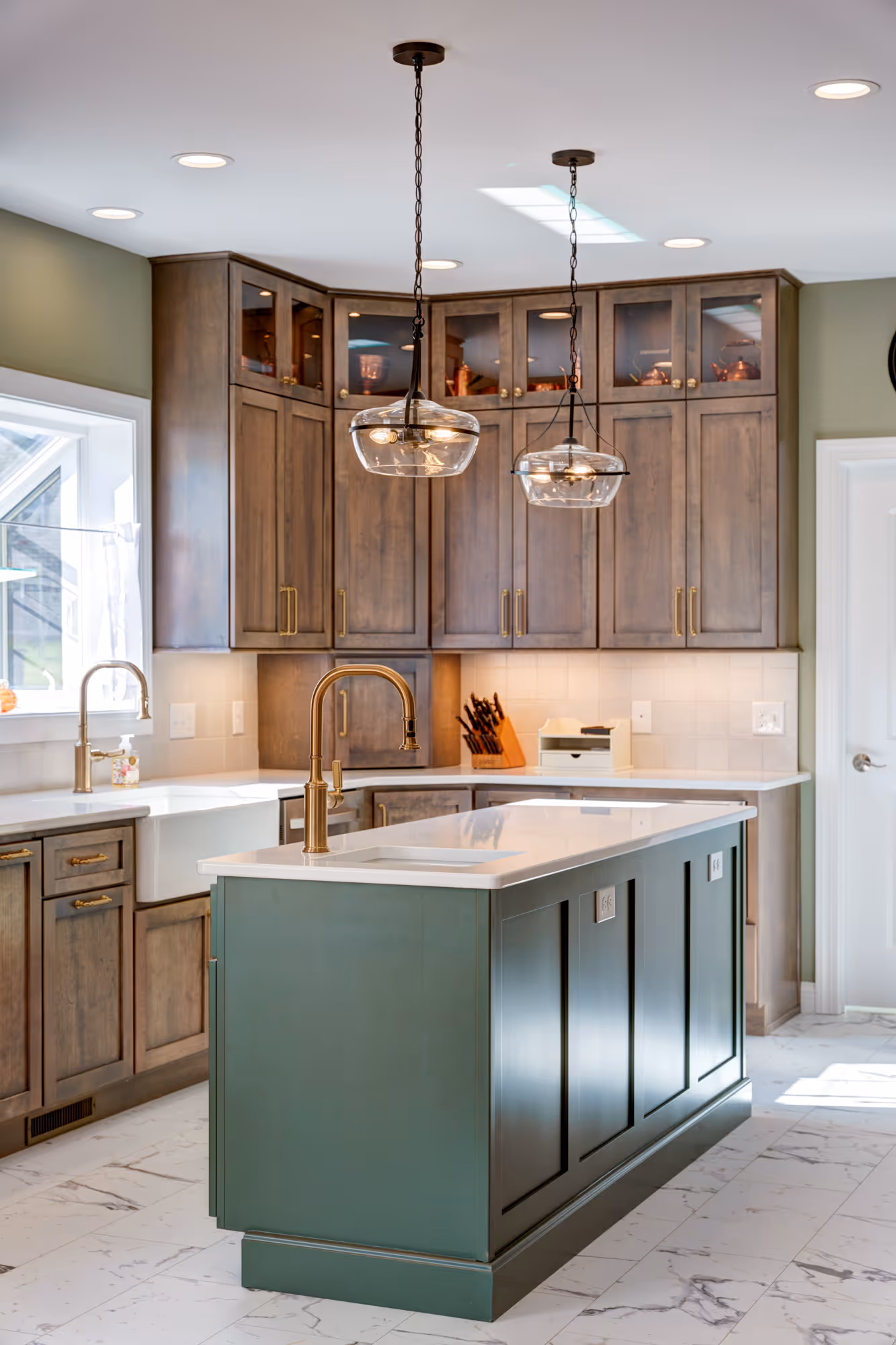 Modern kitchen with green island, wooden cabinets, brass fixtures, and marble floor tiles under pendant lights.