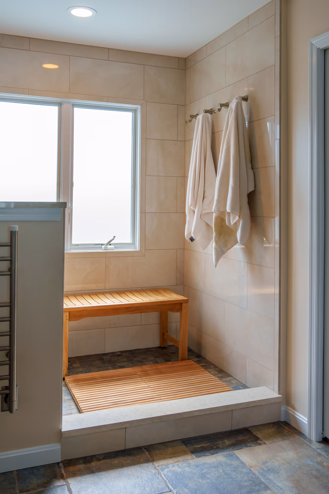 Modern primary bathroom in Mechanicsburg, PA featuring a wooden bench, tiled flooring, and towel holder in a serene shower area.