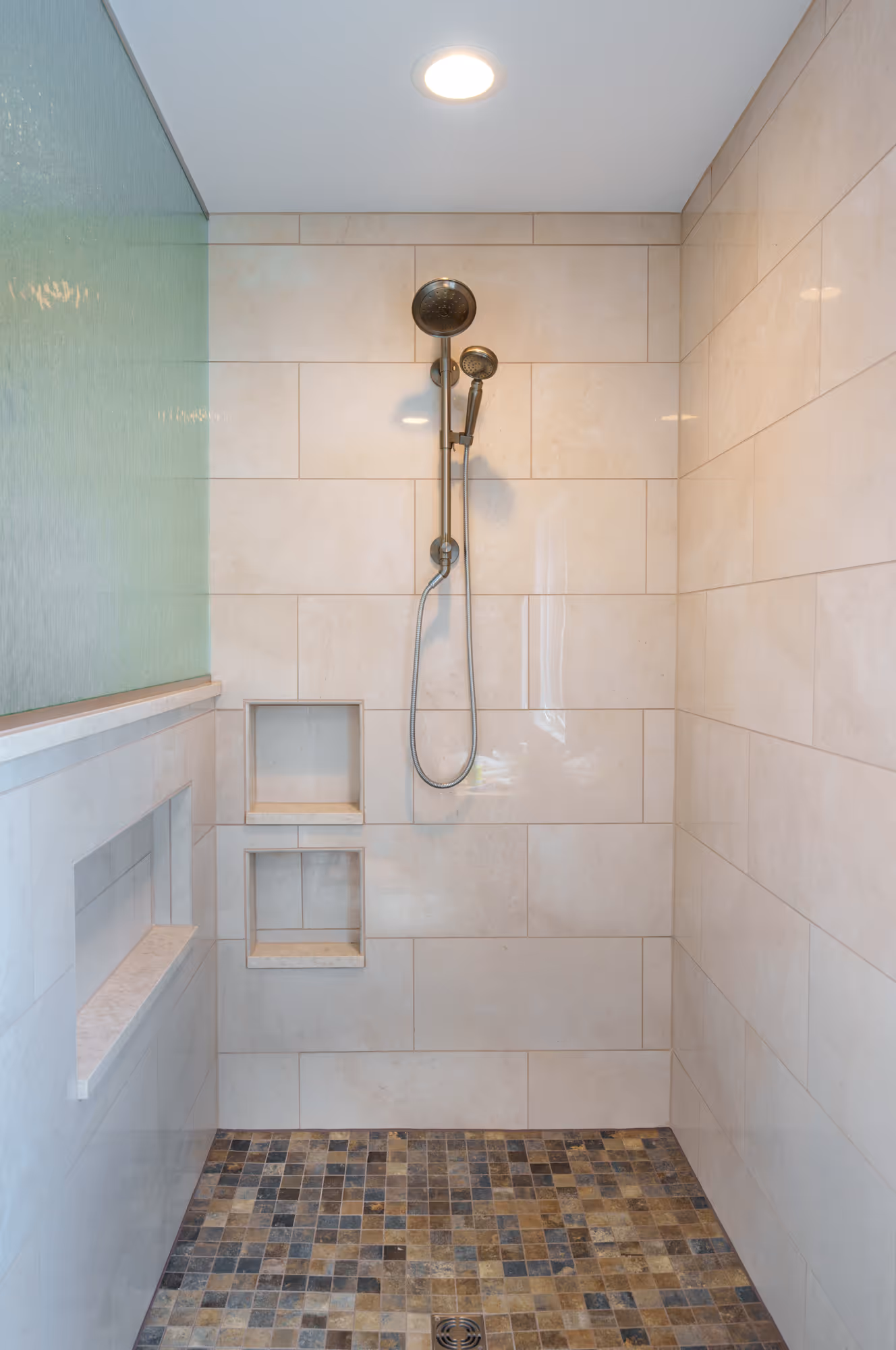Modern primary bathroom shower with beige tiles and a decorative mosaic floor in Mechanicsburg, PA.
