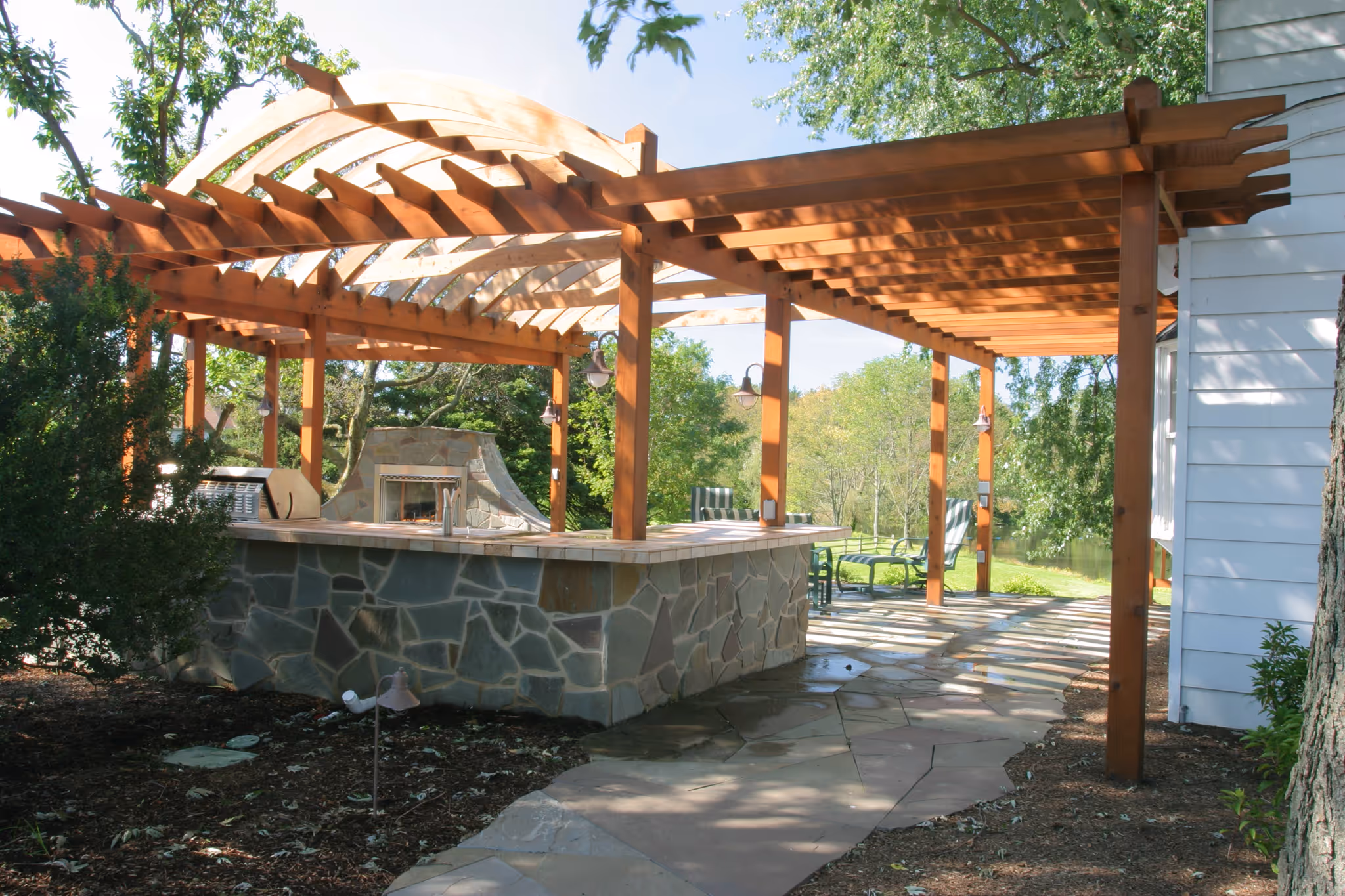Outdoor kitchen with stone accents under wooden pergola in Harrisburg, PA, featuring a fireplace and vibrant greenery