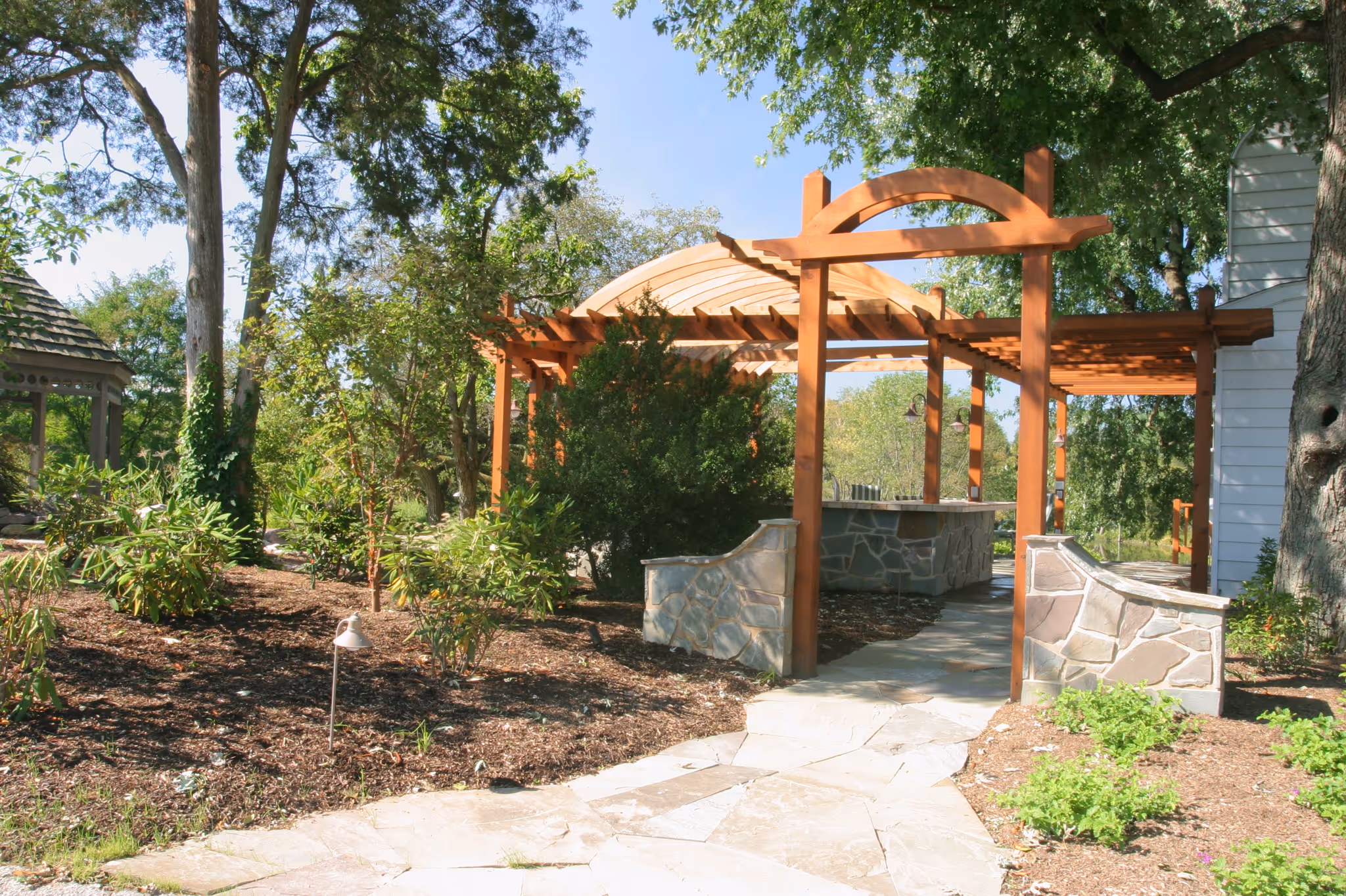 Outdoor pergola with stone seating area in Harrisburg, PA, surrounded by lush greenery and landscaping.
