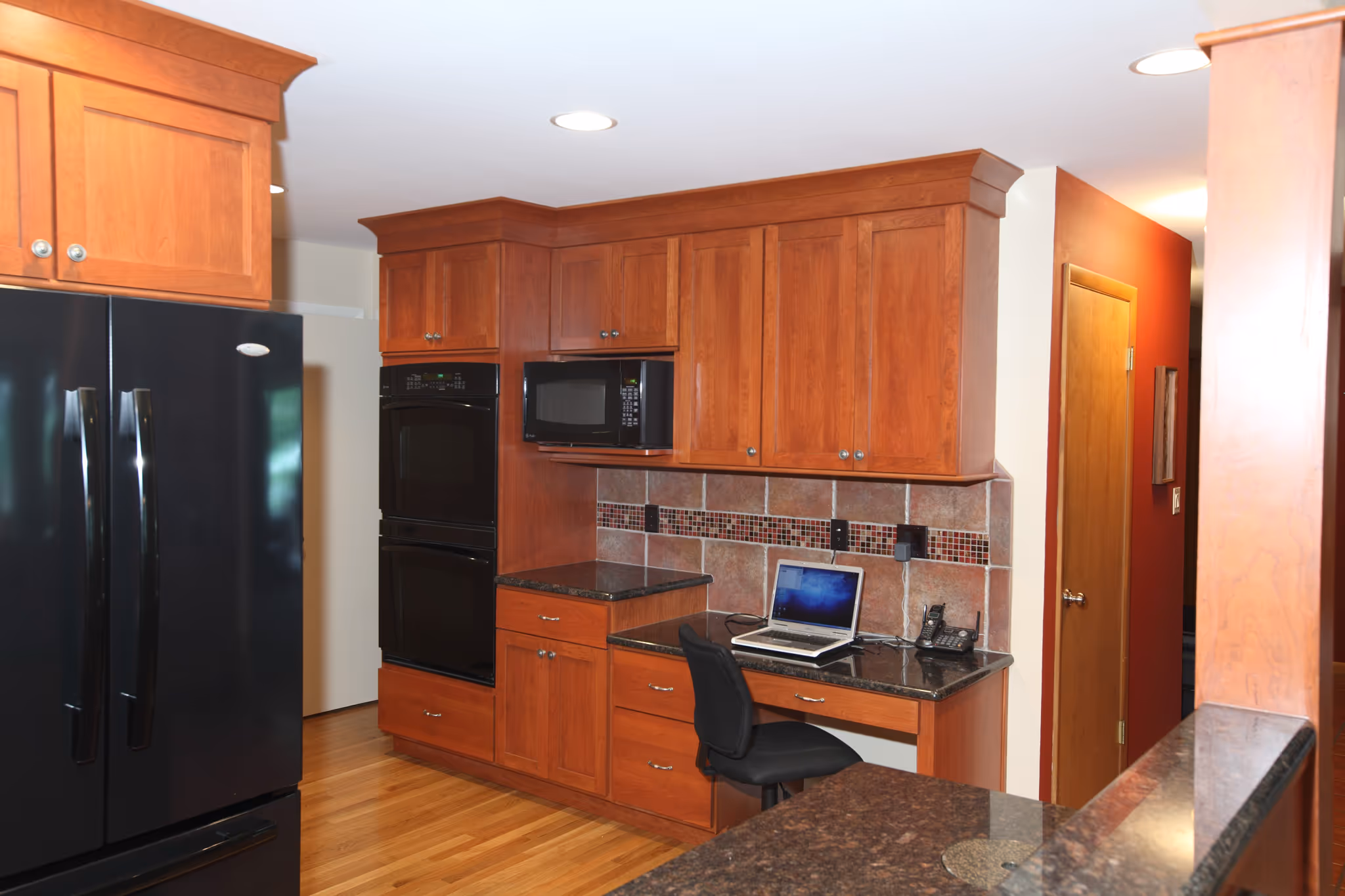 Contemporary kitchen in Carlisle, PA featuring cherry wood cabinets, black appliances, and a desk area with a computer.