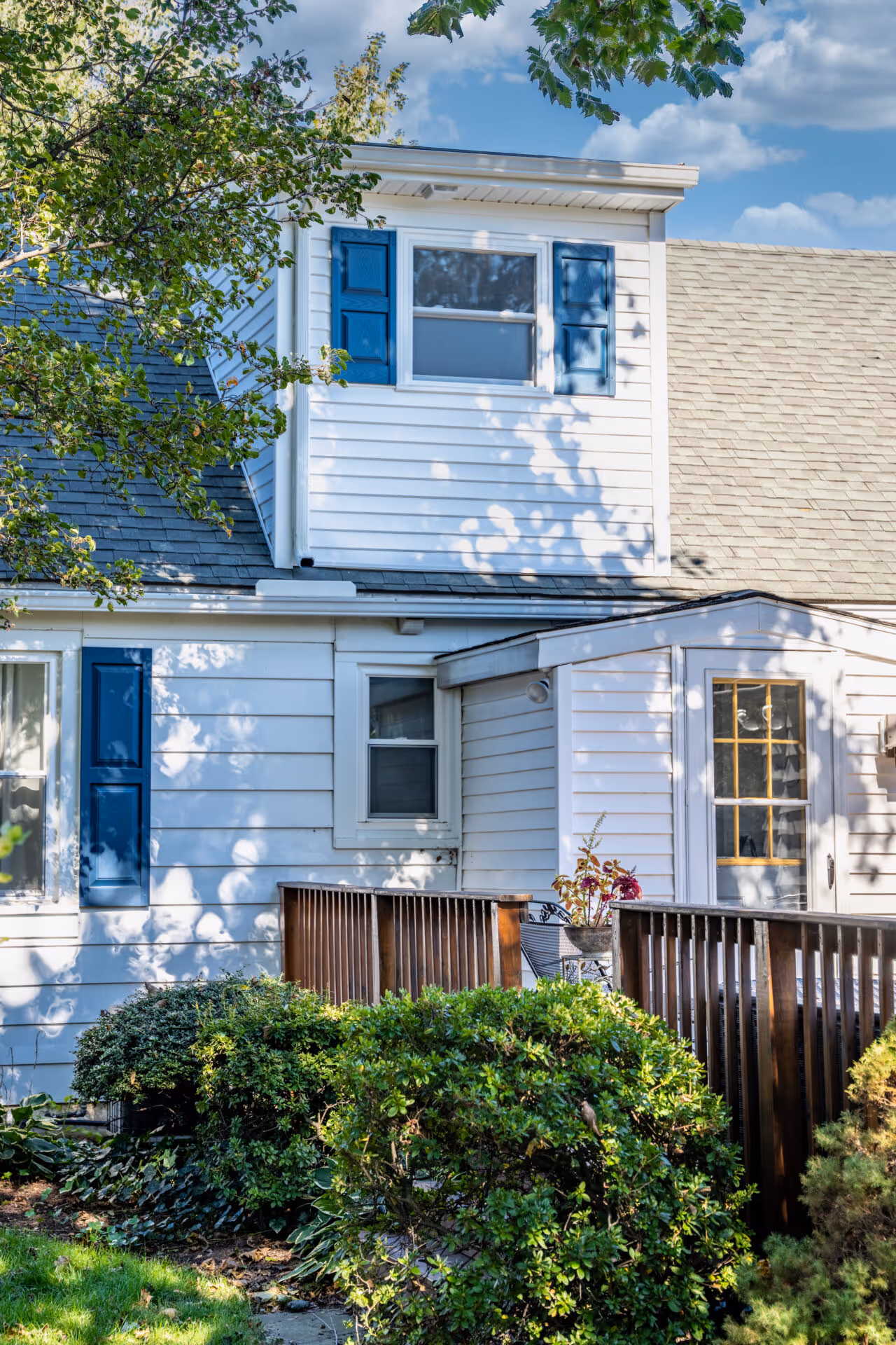 Cozy dormer style exterior with white siding and blue shutters in New Cumberland, PA, featuring a wooden railing and landscaped shrubs.
