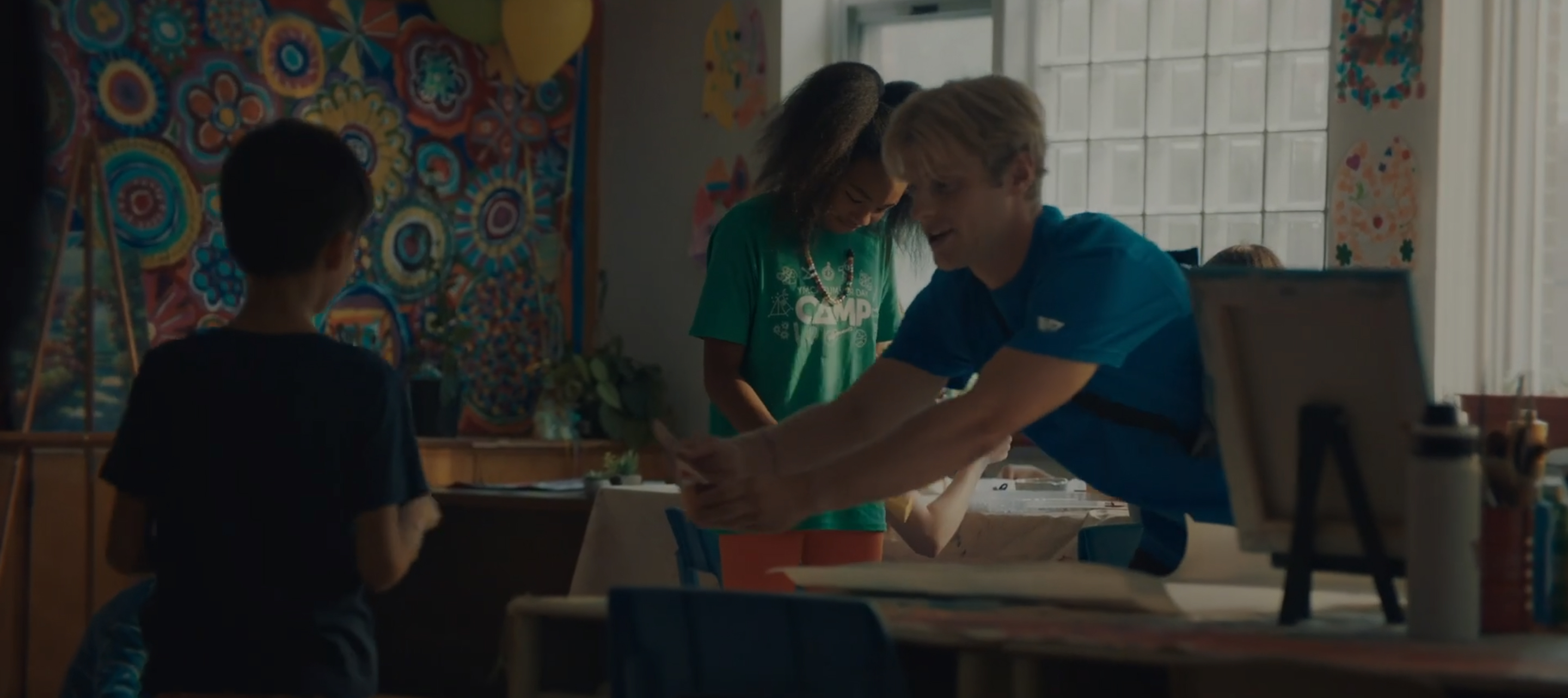 A young man in a blue shirt helps children with art projects in a colorful classroom.