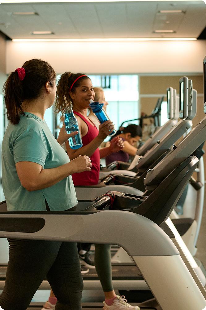 Four women walking on treadmills in a gym, two holding blue water bottles and chatting.