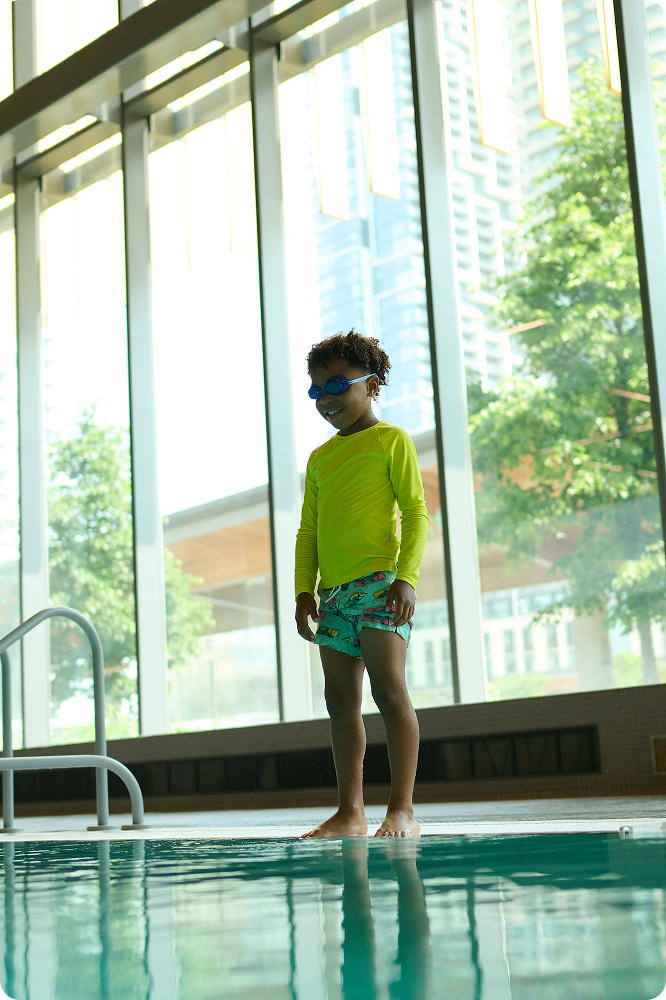 Child wearing goggles and a yellow swim shirt standing at the edge of an indoor pool with large windows behind.