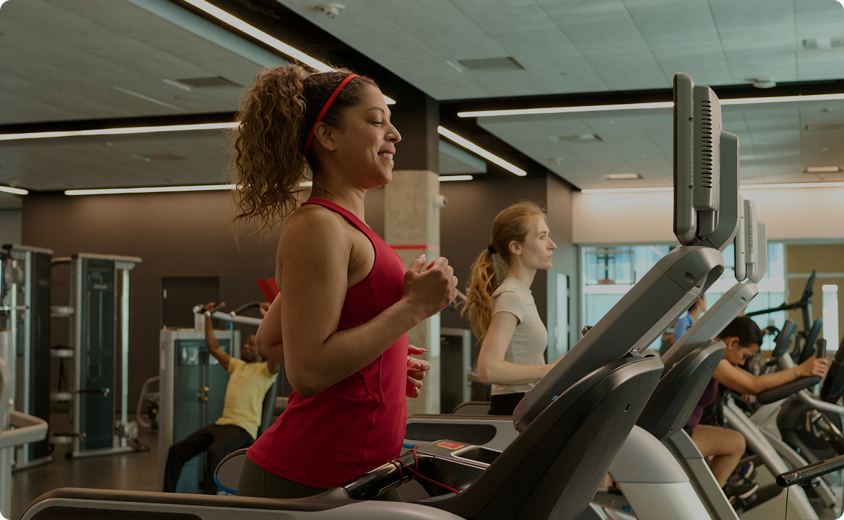 Smiling woman in red tank top running on a treadmill in a gym with other people exercising in the background.
