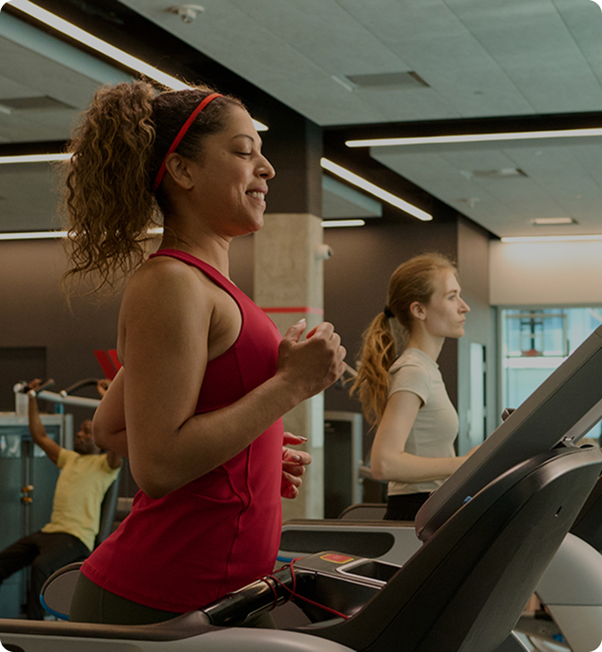 Two women jogging on treadmills in a gym, one wearing a red tank top and headband, the other in a light-colored shirt.