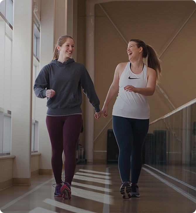 Two women walking hand-in-hand indoors on a track, smiling and dressed in athletic wear.