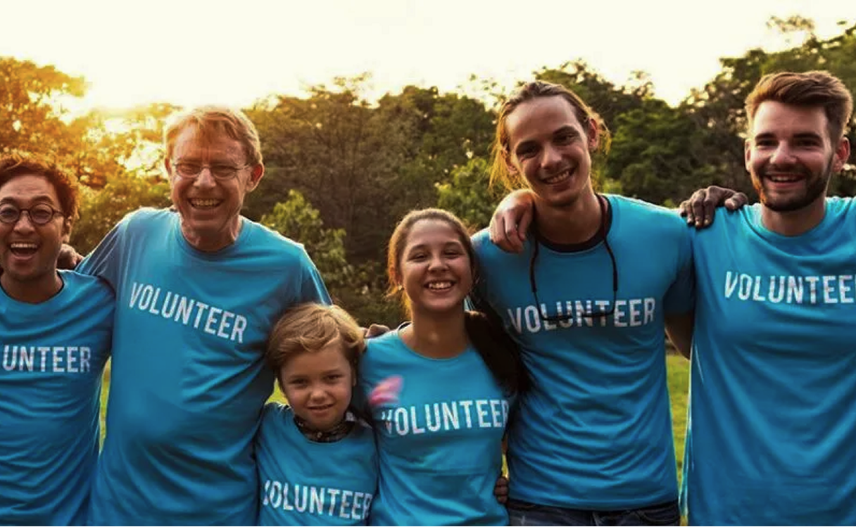 Group of smiling volunteers wearing blue shirts with the word 'VOLUNTEER' outdoors.