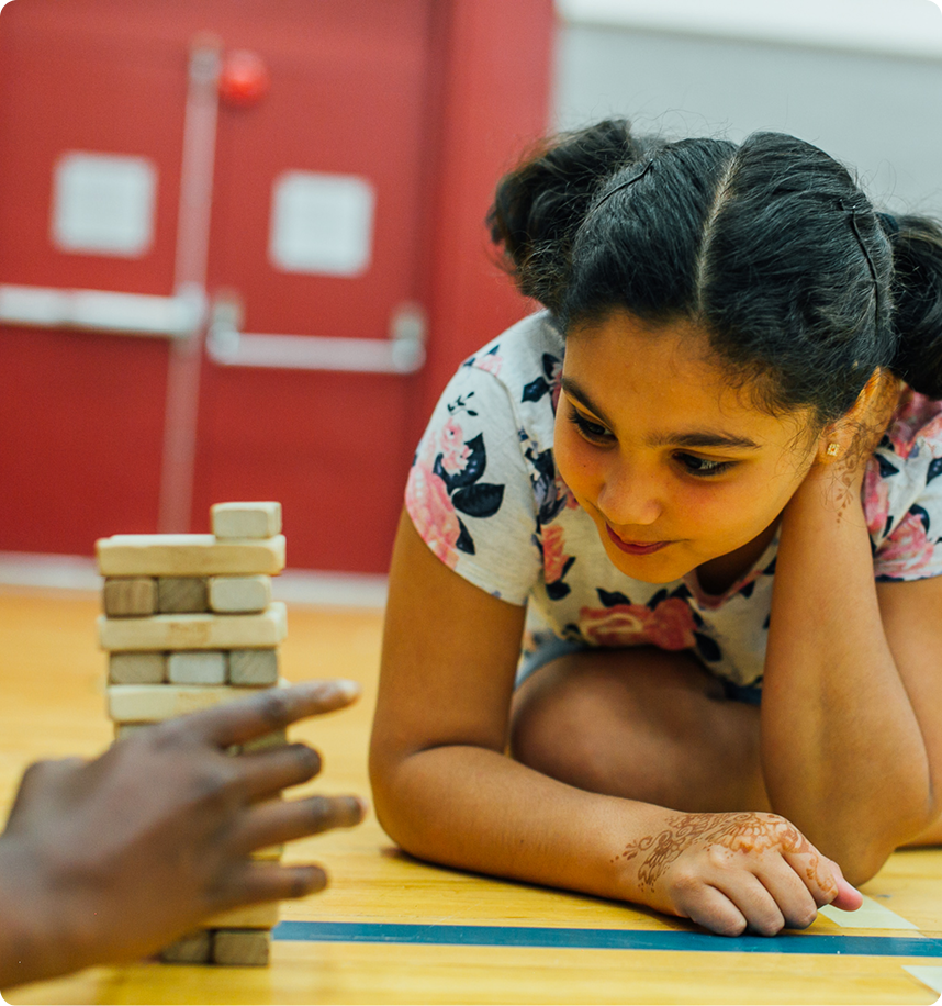 Young girl with henna on her hand watching a hand play Jenga blocks on a wooden floor in a gymnasium.