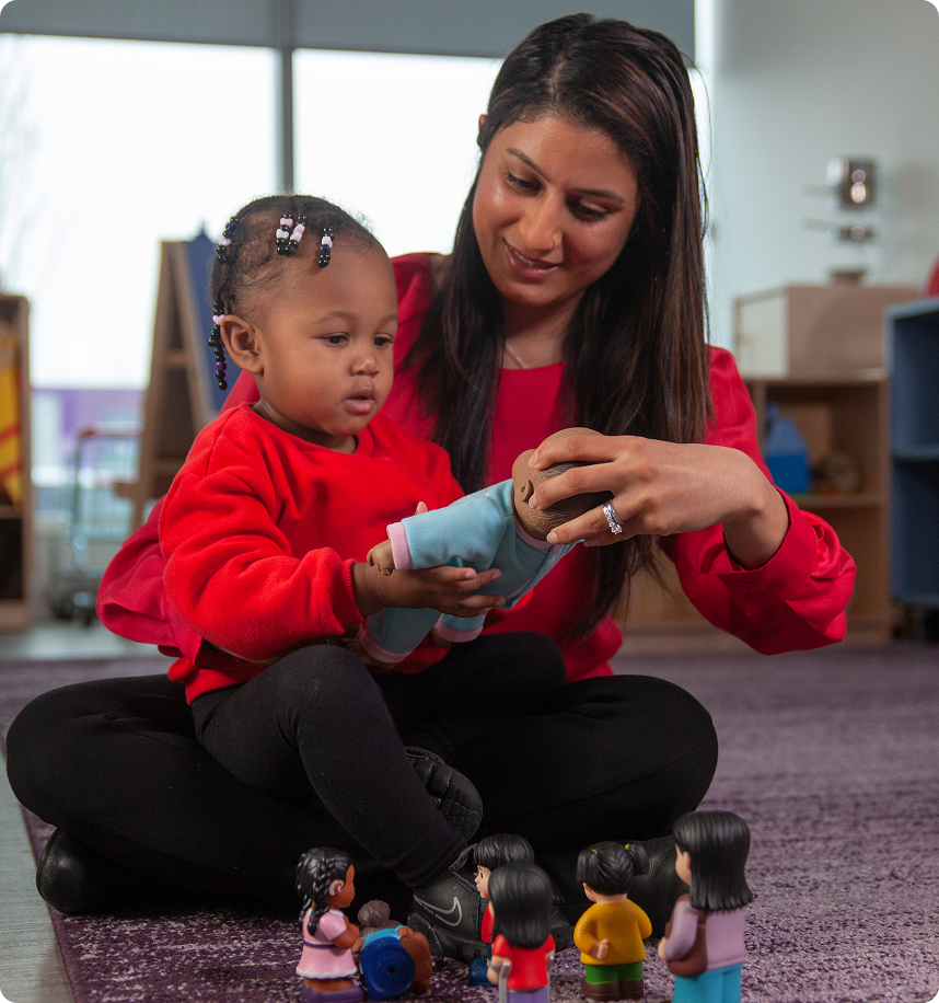 A woman in a red top sits cross-legged on the floor with a young child, showing the child a doll while several small toy figures are arranged in front of them.