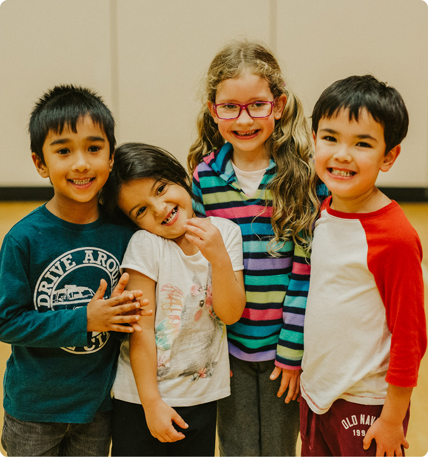 Four smiling children standing close together indoors, two boys and two girls, one girl wearing glasses and a striped sweater.