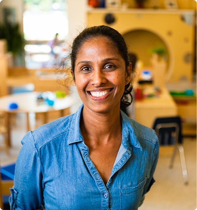 Smiling woman in a denim shirt standing in a colorful classroom environment.