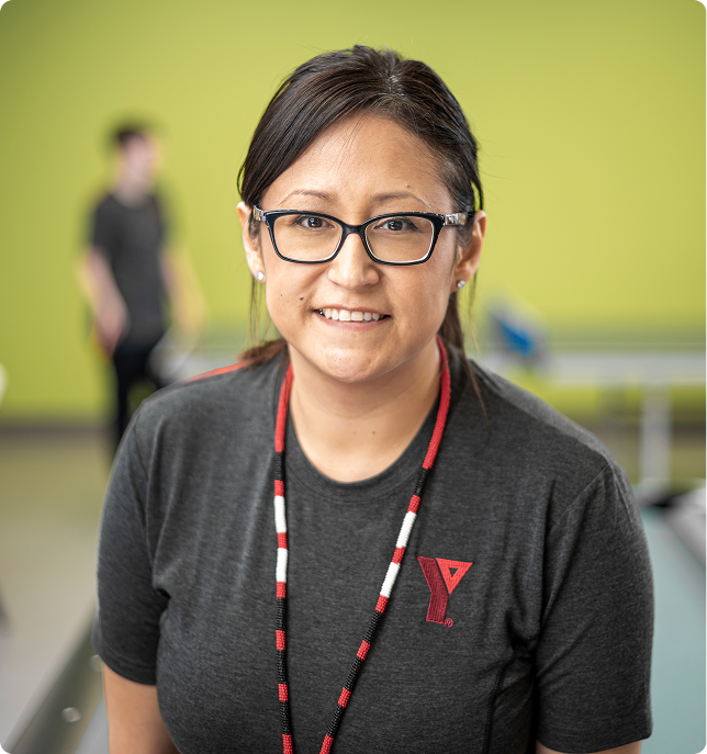 Smiling woman wearing glasses and a dark YMCA shirt with a beaded necklace, standing indoors with a green background.