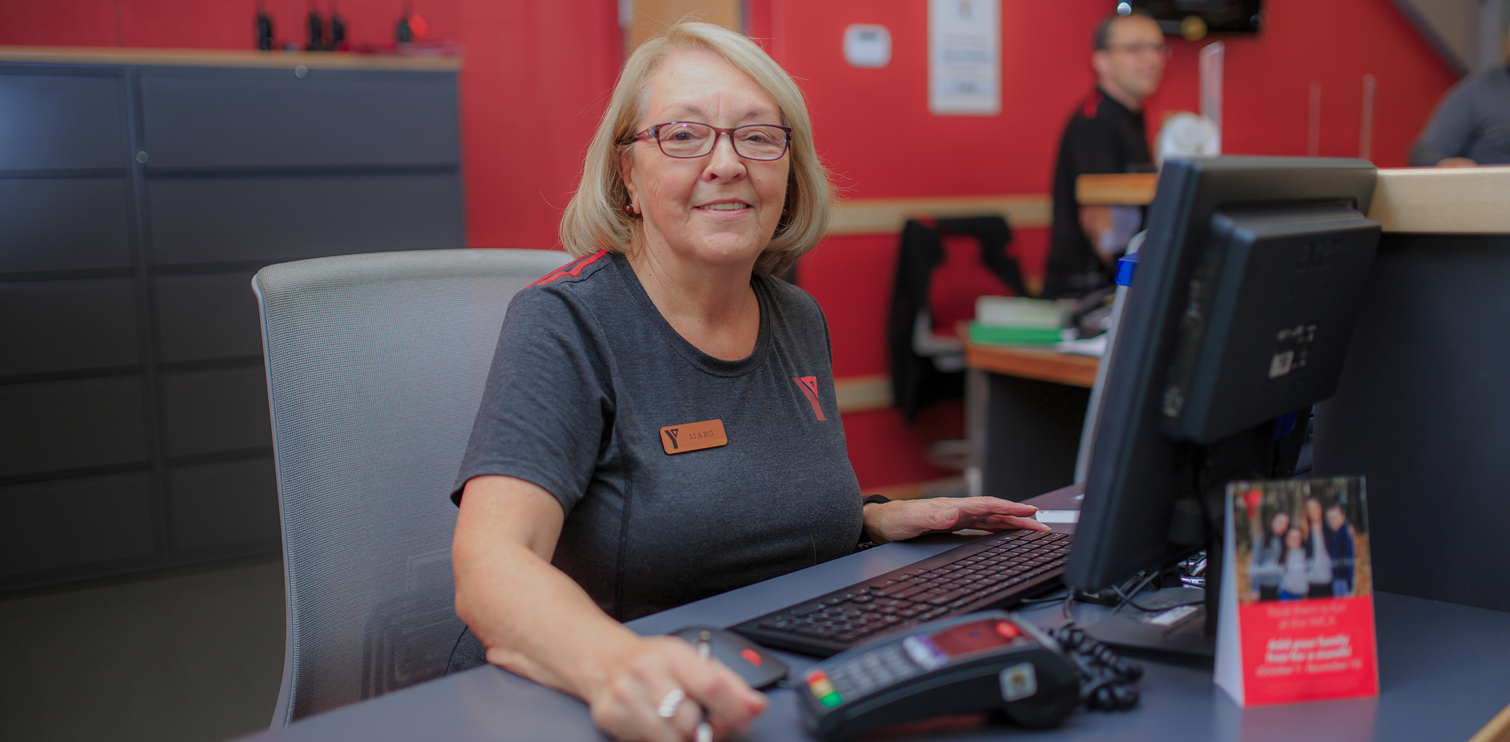 Smiling older woman with glasses and name tag sitting at a reception desk working on a computer.