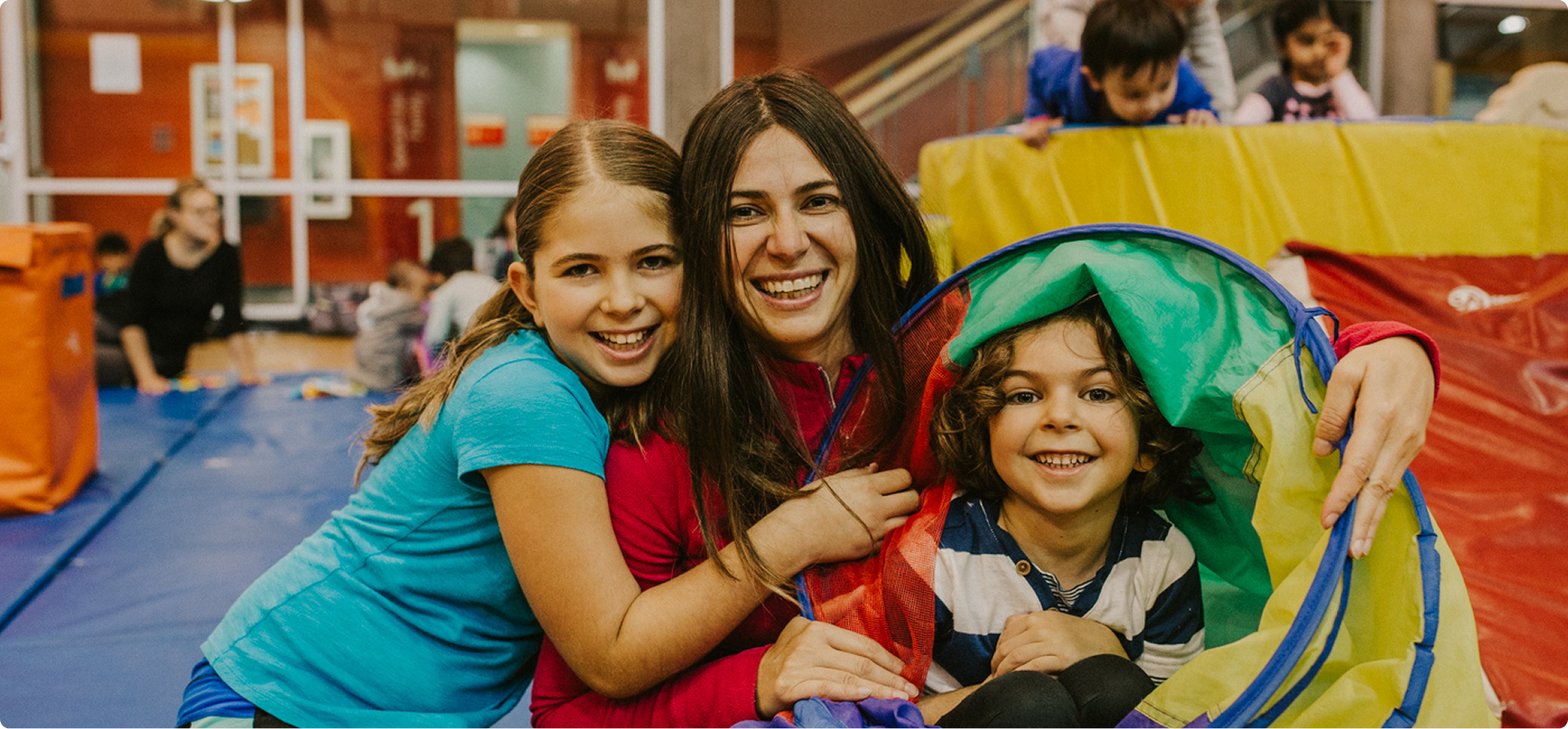 Smiling woman and two children playing in an indoor gym with colorful mats and a play tunnel.