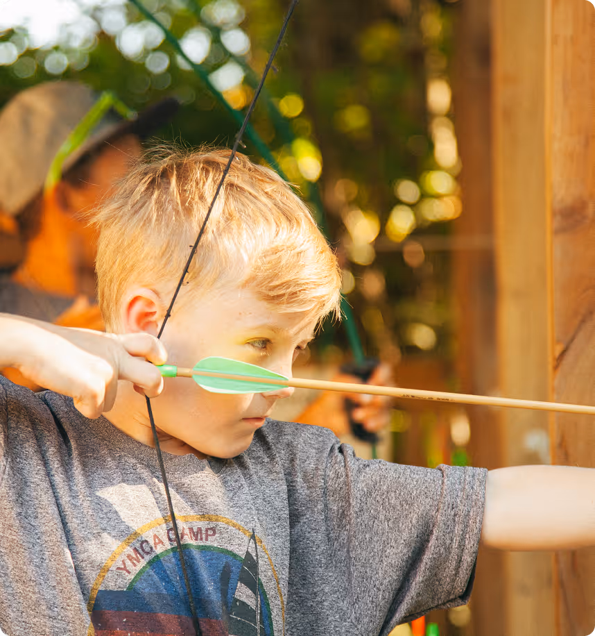 Young boy aiming a green-feathered arrow with a bow during archery practice at an outdoor camp.