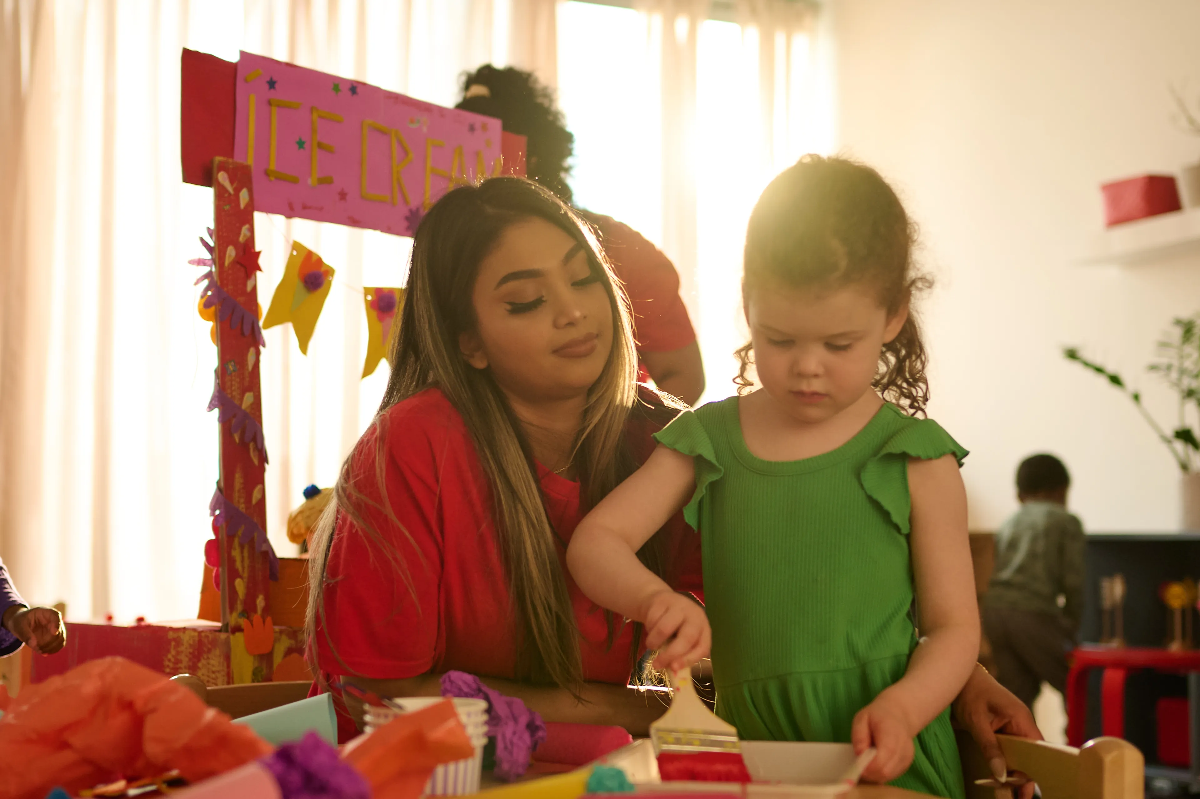 Young woman in red shirt watching a little girl in a green dress paint with a brush at a colorful craft table.