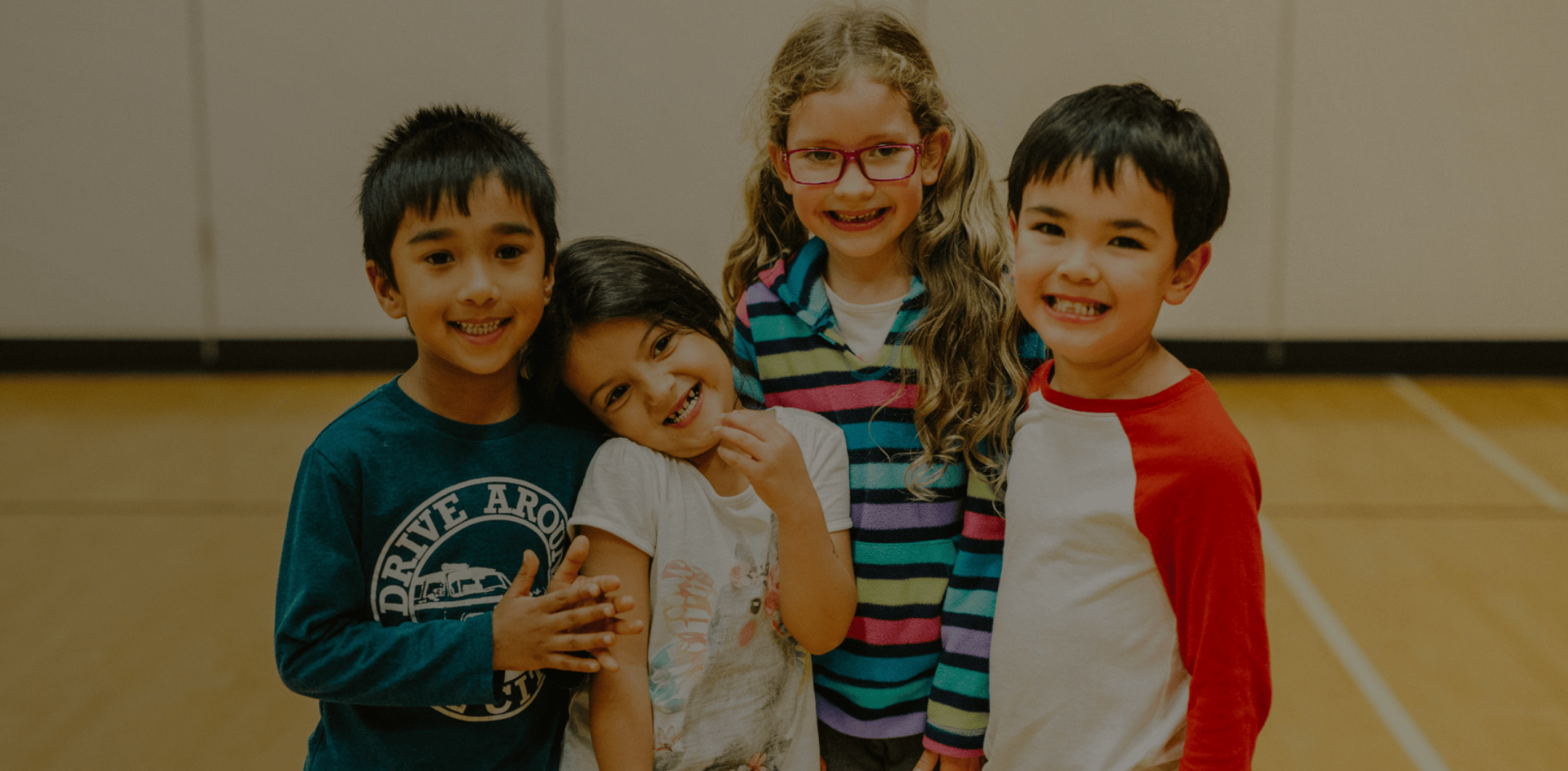 Four smiling children standing closely together indoors on a gym floor, two boys and two girls, one girl wearing glasses and a colorful striped sweater.