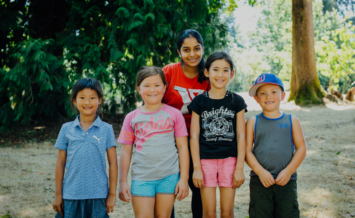 Smiling group of one young woman and four children standing outdoors on grass with trees in the background.
