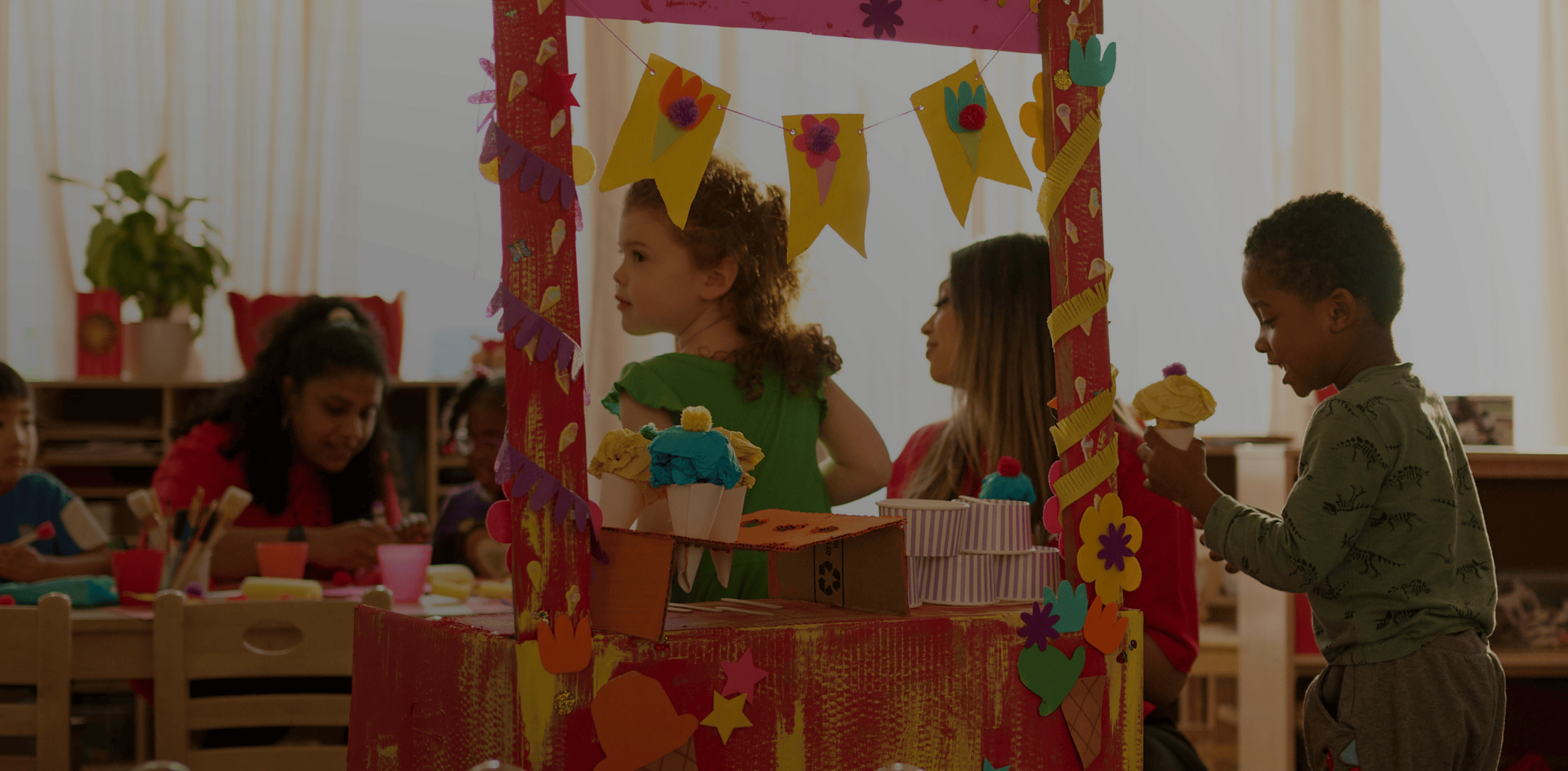 Children playing and crafting colorful paper ice cream cones at a decorated arts and crafts table in a classroom.
