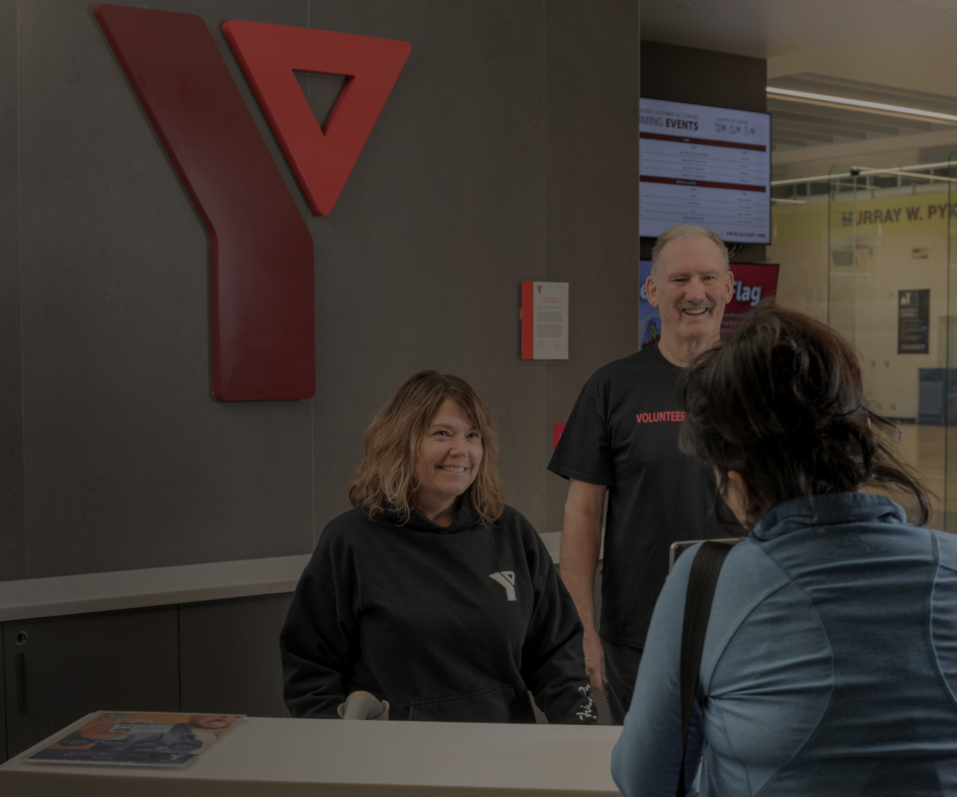 Two YMCA staff members smiling behind a reception desk while a visitor talks to them.