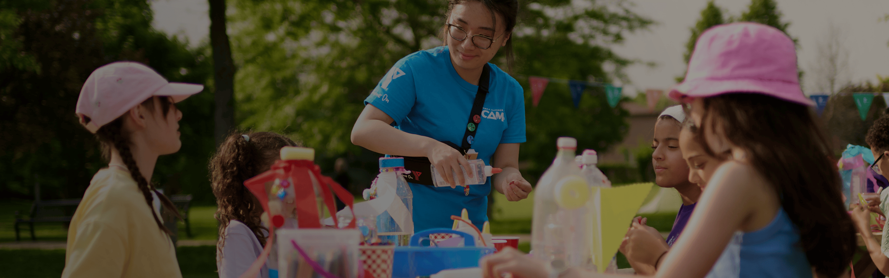 Children and a counselor engaging in crafts around picnic tables outdoors at a YMCA day camp.