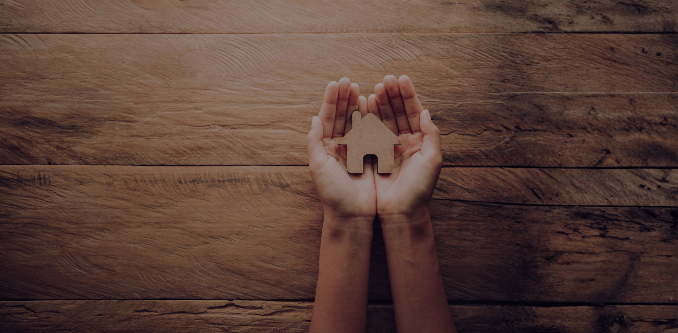 Two hands gently holding a small wooden house cutout over a wooden surface.