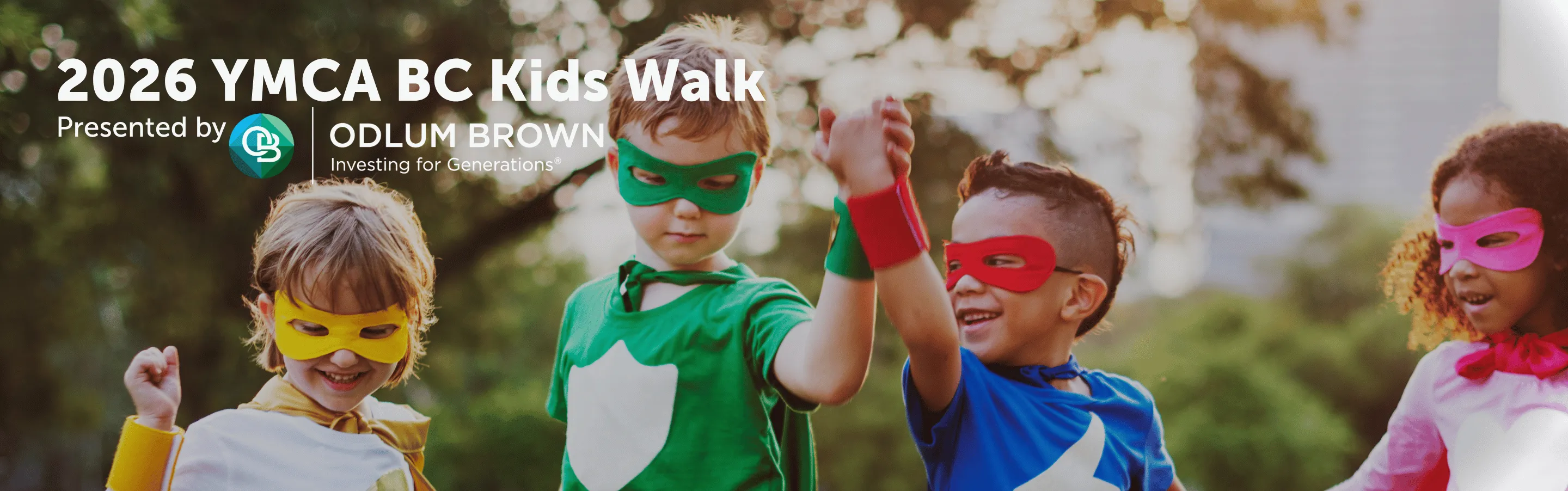 Four children dressed as superheroes with colorful masks and capes holding hands outdoors.
