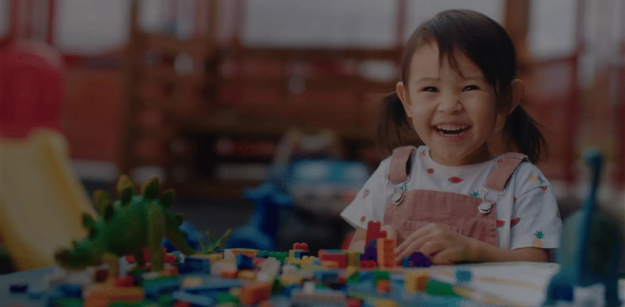 A young girl with pigtails smiling while playing with colorful building blocks and toy dinosaurs on a table.