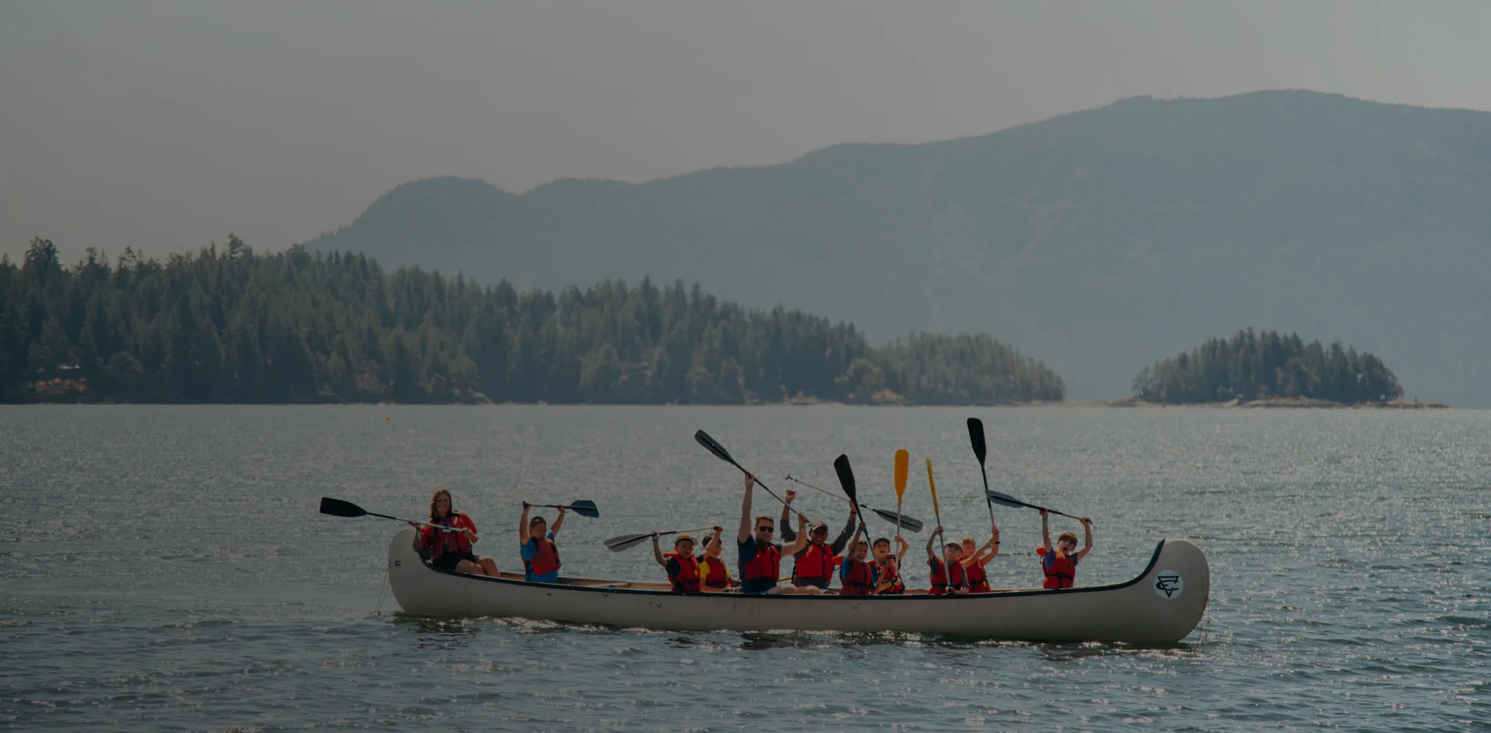 Group of people in life jackets raising paddles while canoeing on a lake with forested hills in background.