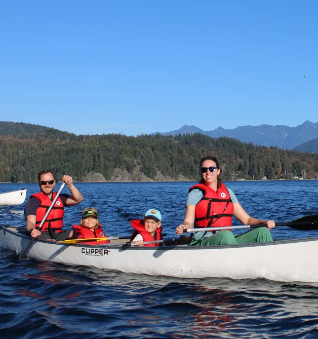 Family of four wearing life jackets paddling a canoe on a lake with forested hills behind them.