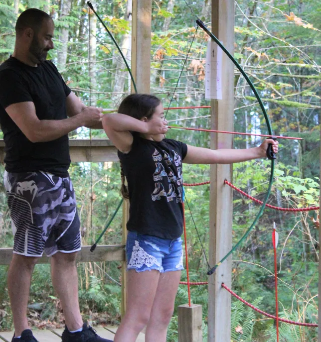 Man assisting girl aiming a bow and arrow at an outdoor archery range surrounded by trees.