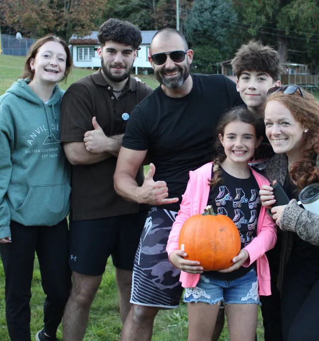 Group of six people smiling outdoors, with a young girl holding a pumpkin in the front.