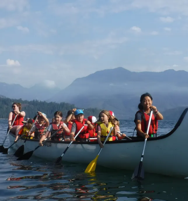 Group of children and adults wearing life jackets paddling a canoe on calm lake with mountains in background.