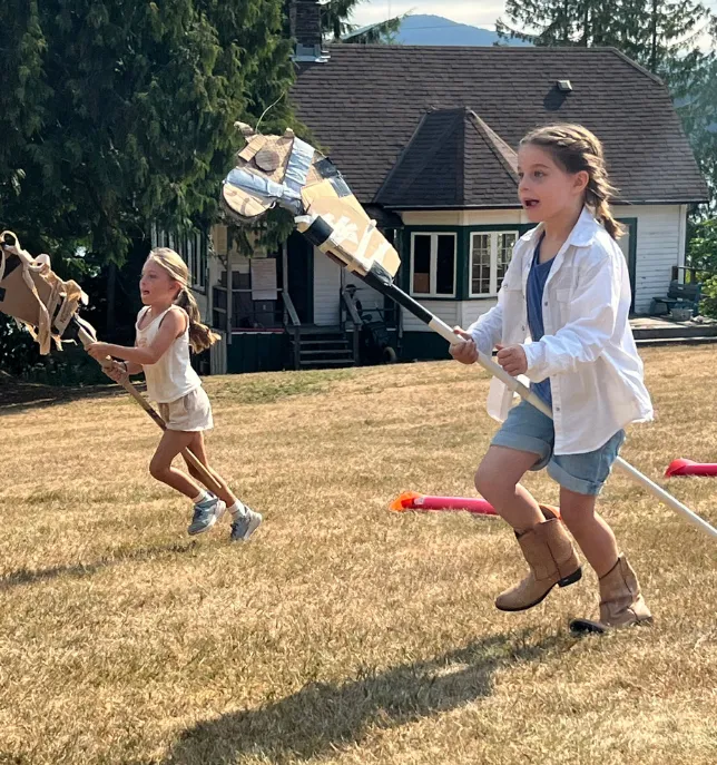 Two girls running on dry grass holding DIY stick horses near a house on a sunny day.