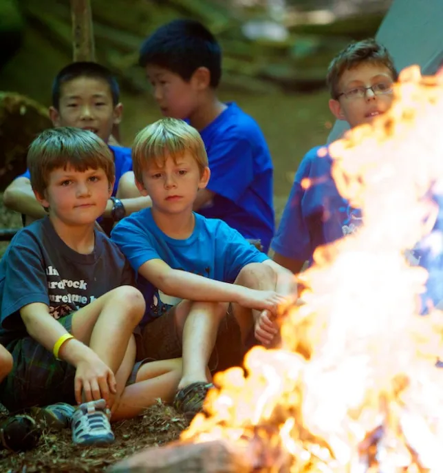 Group of boys sitting near a bright campfire outdoors in a wooded area.