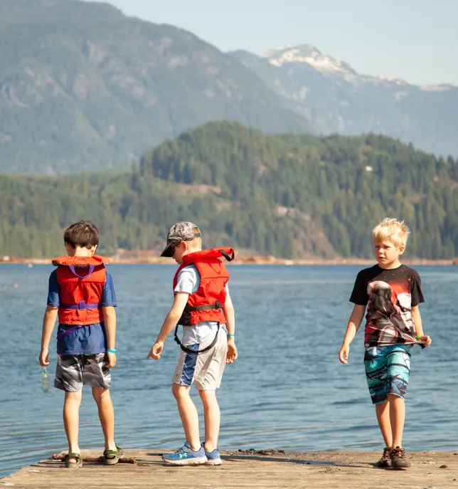 Three boys on a dock by a lake, two wearing red life jackets, with forested mountains in the background.