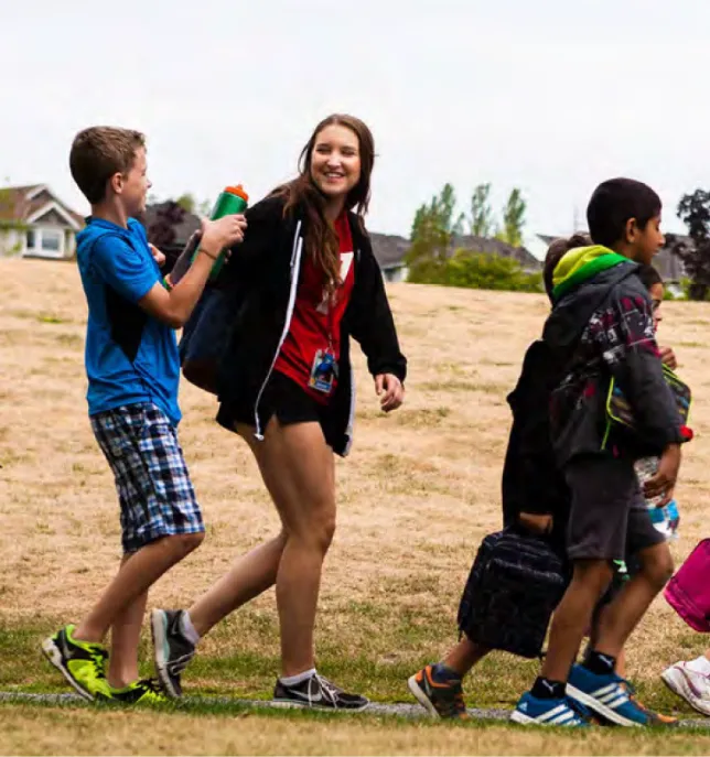 Smiling young woman walking with kids on grass, one kid holding out a green water bottle.