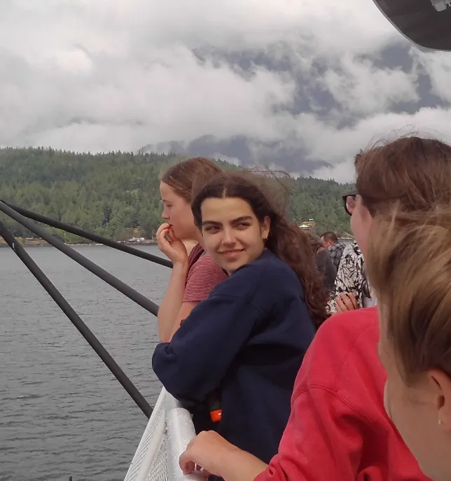 Group of young people leaning on a boat railing looking at water and forested shore under cloudy sky.