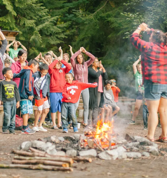 Children and adults standing near a campfire in forest, copying instructor's hand motions outdoors.