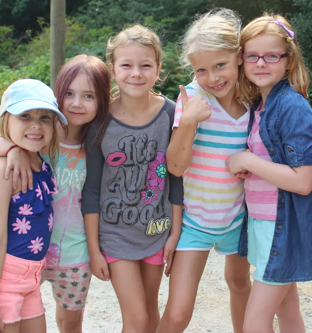 Five smiling girls standing close outdoors, one giving a thumbs-up, wearing casual summer clothes.