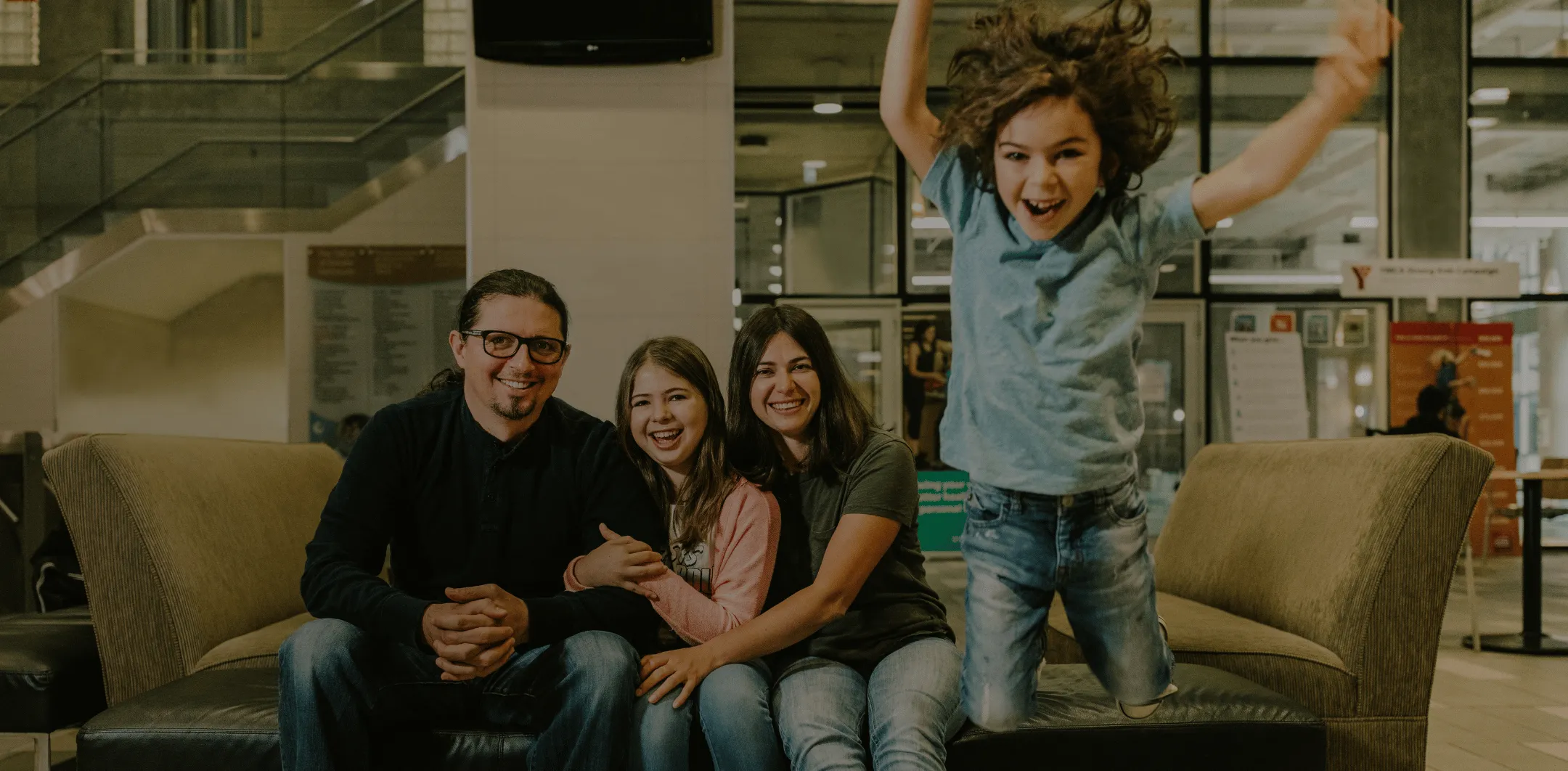 Smiling family of three sitting on a couch while a child joyfully jumps beside them in a public indoor space.