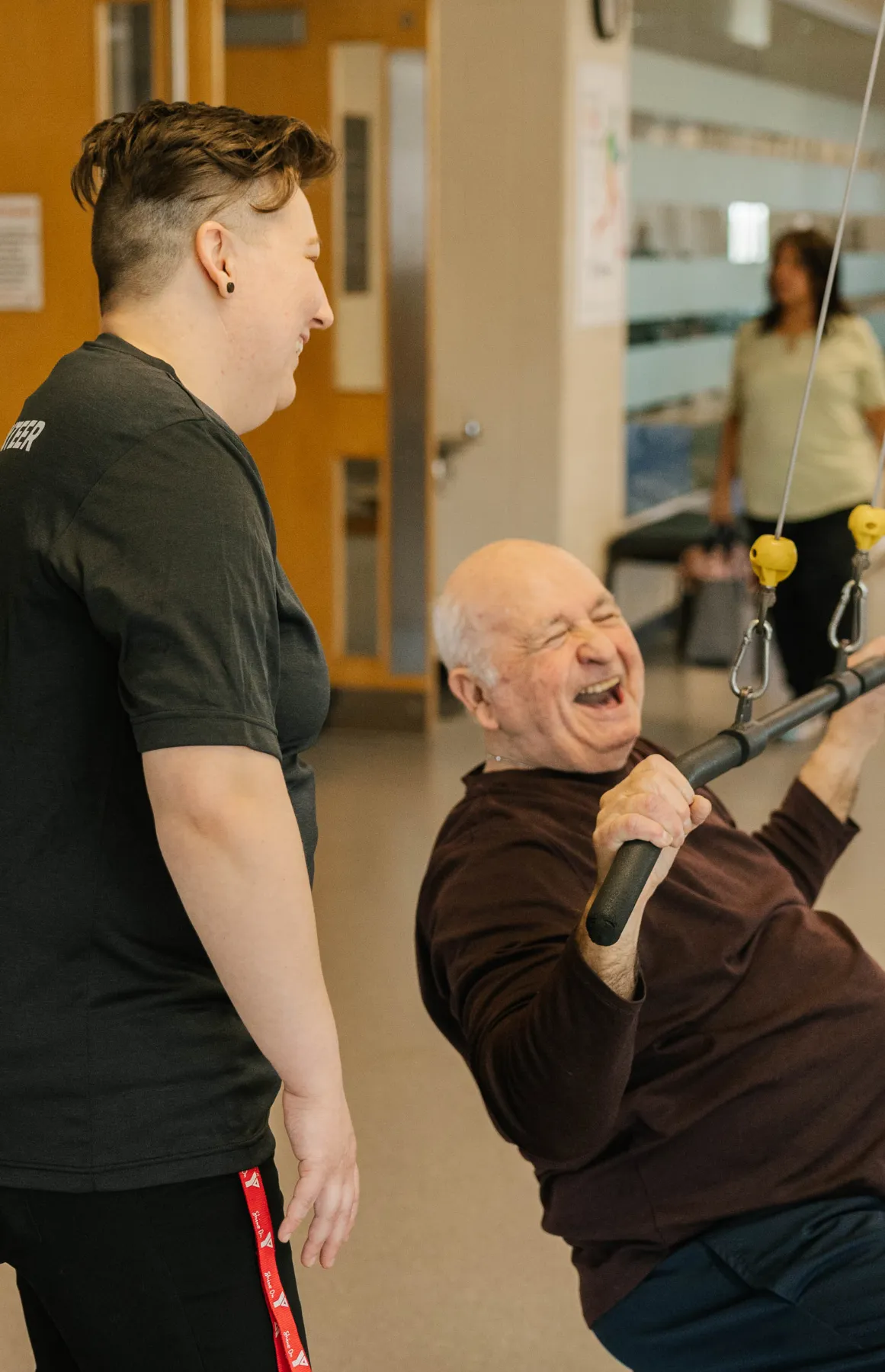 Older man laughing while using a cable exercise machine, assisted by a smiling trainer in a gym.